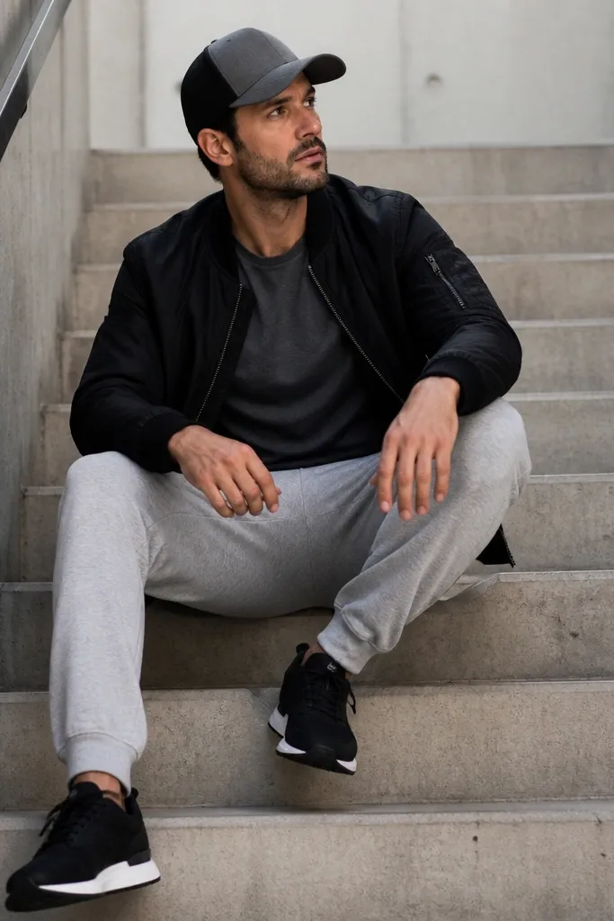 Grey sweatpants with a black bomber jacket. The man wears a black-and-grey cap, a dark t-shirt, and black sneakers with white midsoles. The photo is taken on a staircase.