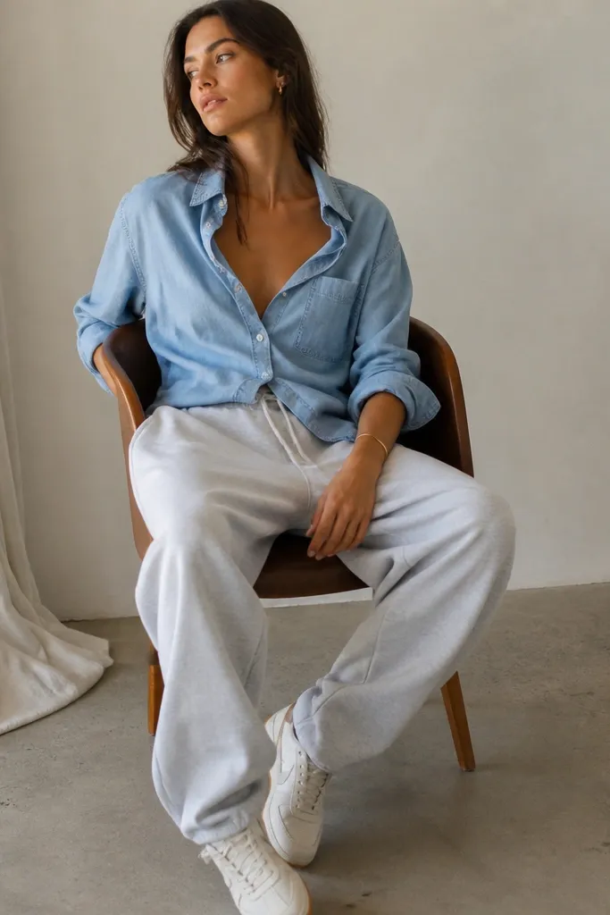 A model wearing baggy grey sweatpants with a light blue chambray button-up. The shirt is worn with the top buttons open and sleeves rolled slightly. White retro sneakers and a simple bracelet.