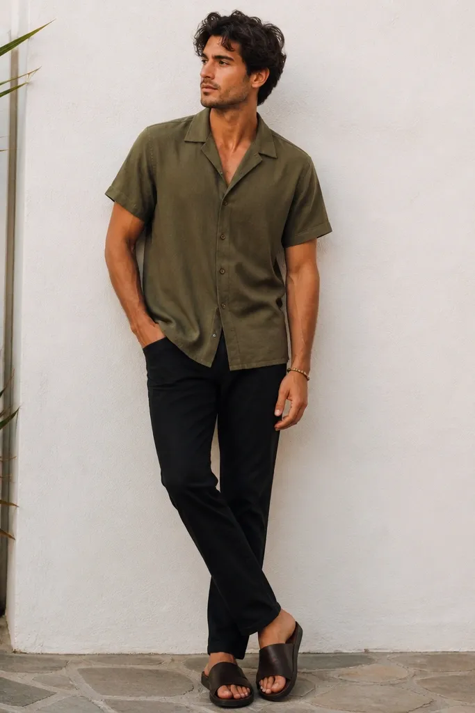 Black jeans with an olive camp collar short-sleeve shirt, collar open, shirt untucked, and dark brown slide sandals; a woven bracelet on the wrist.