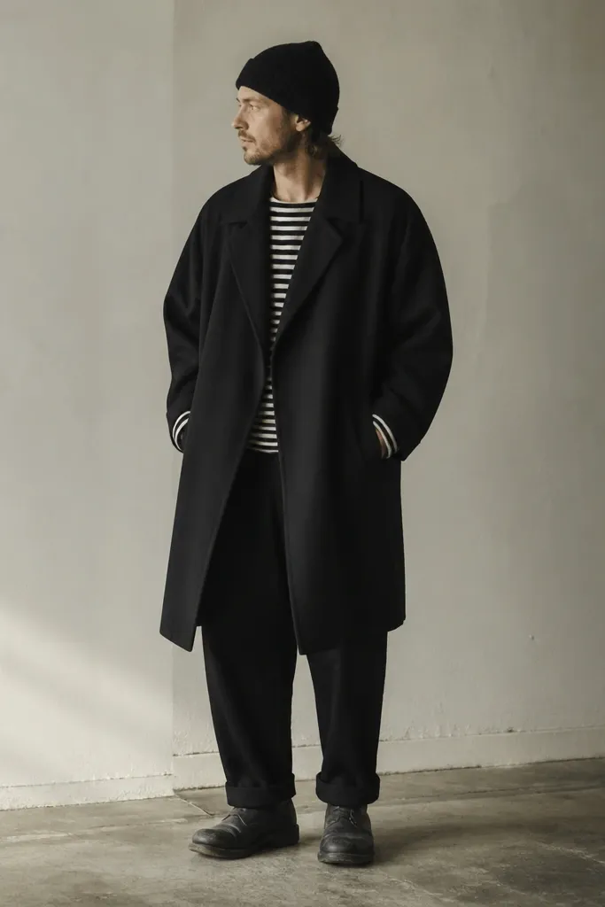 Man in an oversized black coat worn open, a black-and-white striped thermal shirt peeking at the neck and cuffs, black pants, and worn black boots. A chunky knit beanie covers his head.