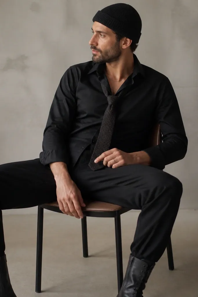 Man wearing a black button-up shirt slightly open at the collar, a knit tie in dark gray, black pants, and combat boots. He has a chain necklace and a beanie.