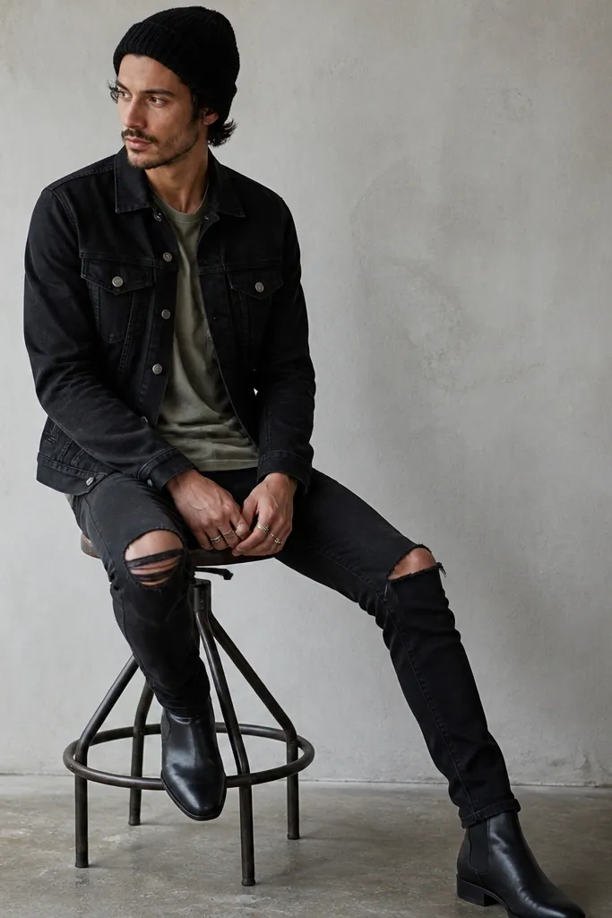 A man wearing a faded olive T-shirt under a black denim jacket with metal buttons. He has black ripped knee jeans with frayed edges and a narrow silhouette. Shoes are black leather boots with a slight heel, and he wears a knit cap.