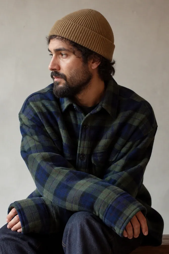 A man in a green-and-navy plaid flannel with sleeves that extend past his hands, bunching at the wrist. He's wearing a tan knit beanie and dark jeans. The frame shows the cuff covering part of the palm.