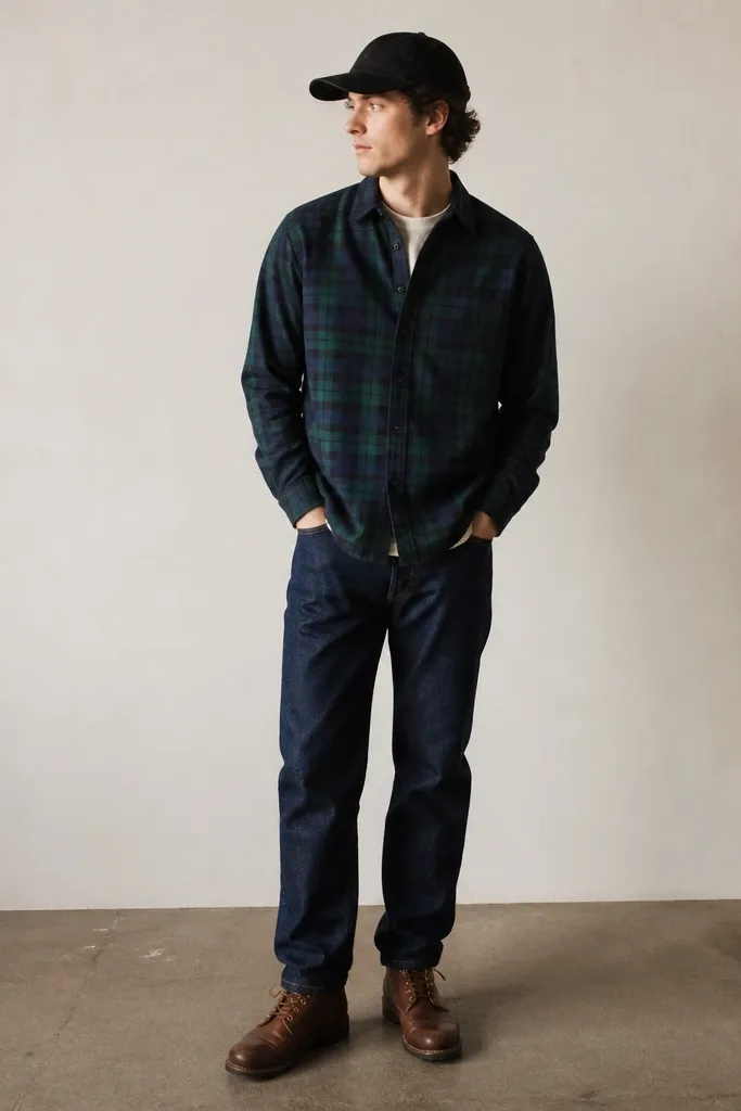A model wearing a blackwatch plaid flannel in deep green and navy, buttoned up over an off-white crewneck tee. Straight-leg dark indigo jeans sit at the ankle with a slight break. Brown leather lace-up boots and a black baseball cap complete the outfit.