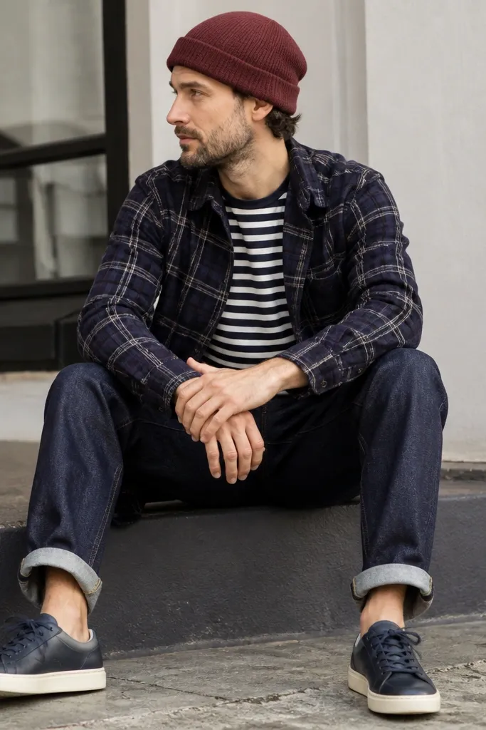 A navy plaid flannel worn open over a navy-and-white striped tee. Indigo selvedge jeans with visible stitching at the cuff. Leather sneakers in dark navy and a maroon beanie.